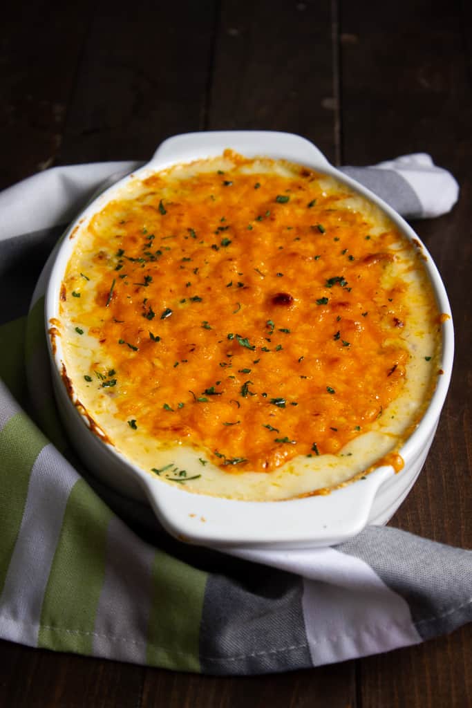 A white oval baking dish filled with baked chicken and cheese macaroni, topped with melted golden-brown cheese and sprinkled with herbs, sits on a striped green, white, and gray kitchen towel on a dark wooden surface.