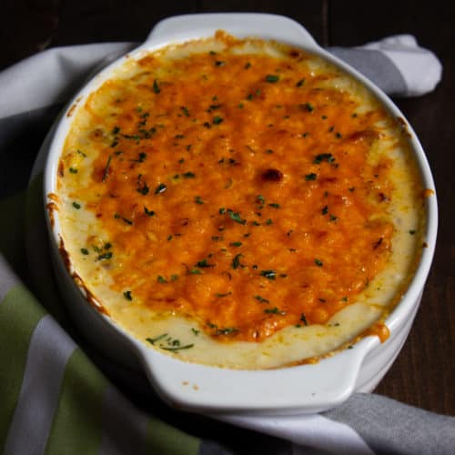 An oval white baking dish filled with a cheesy, golden-brown Chicken and Cheese casserole sits on a green, white, and gray striped cloth, atop a dark wooden surface.