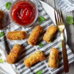 A plate with crispy Garlicky Potato & Spinach Croquettes and a small bowl of ketchup on newspaper, garnished with cilantro, a gold fork, and a glass of water on a wooden table.