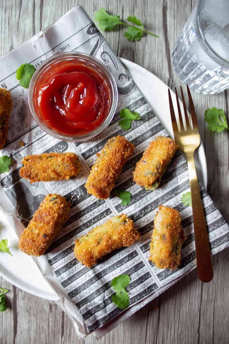 A plate with crispy Garlicky Potato & Spinach Croquettes and a small bowl of ketchup on newspaper, garnished with cilantro, a gold fork, and a glass of water on a wooden table.