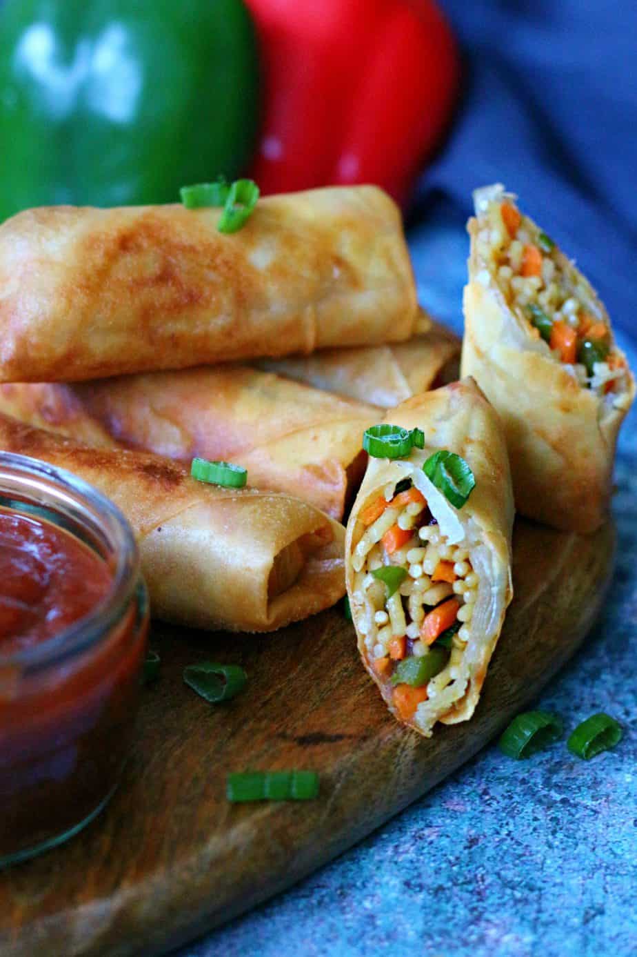 Crispy Vegetable Noodle Spring Rolls on a wooden board, some whole and some cut open to reveal noodles and veggies inside. Garnished with chopped green onions and served with a small jar of dipping sauce.