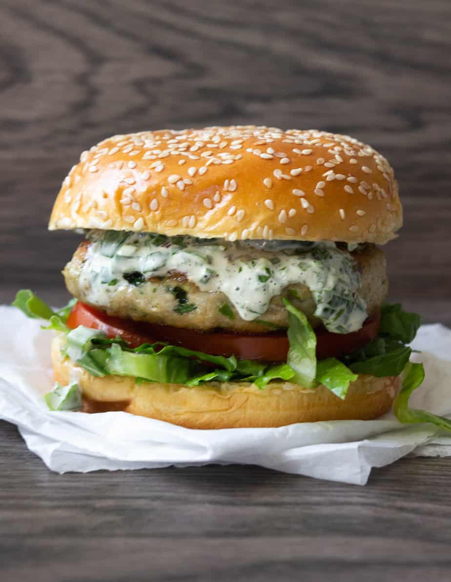 An Herbed Chicken Burger with lettuce, tomato, and creamy herb sauce on a sesame seed bun, resting on white parchment paper against a rustic wooden background.