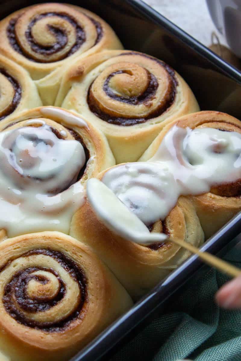 A close-up of Soft Cinnamon Rolls in a pan, with icing being spread on top of the freshly baked rolls using a spoon.