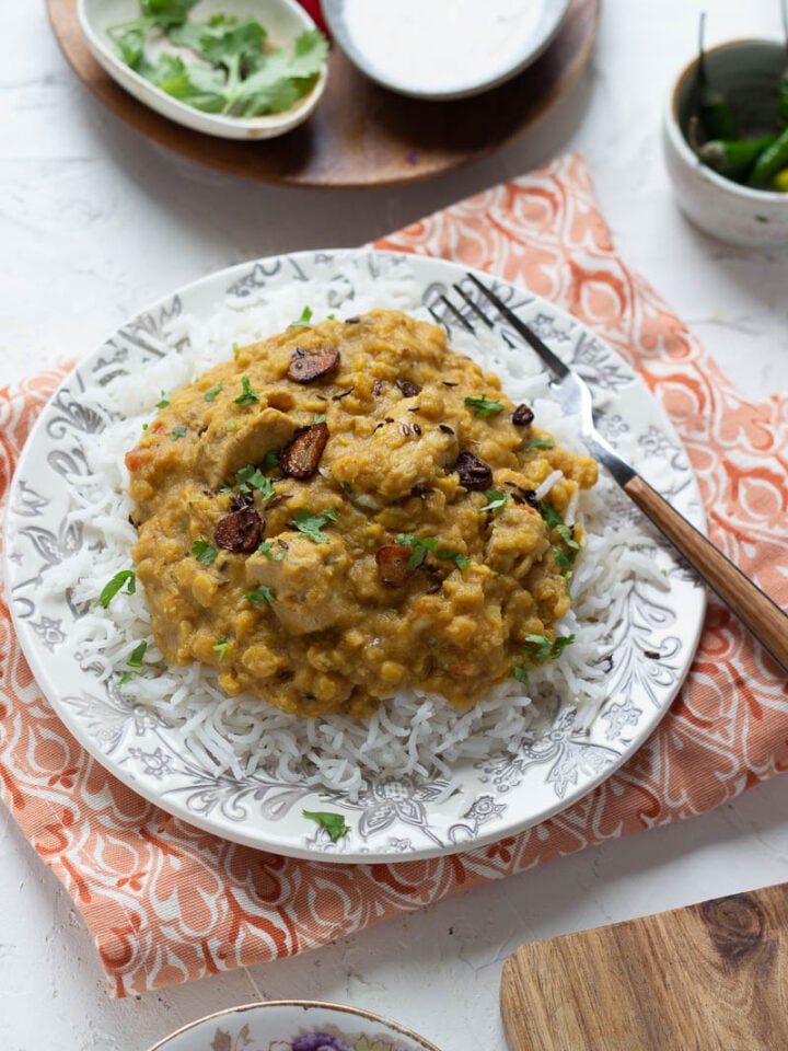 A plate of rice topped with creamy yellow dal chicken, garnished with fried garlic and cilantro. A fork rests on the plate, with small bowls of herbs, sauce, and chilies in the background.
