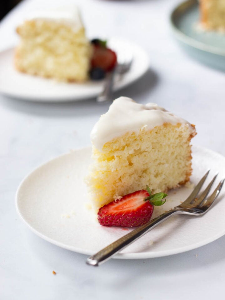 A slice of Lemon Cake with Cream Cheese frosting sits on a white plate, accompanied by a halved strawberry and a fork. Another slice of cake is blurred in the background.