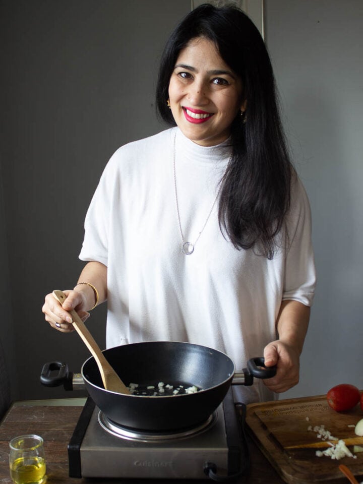 A woman smiles while stirring chopped onions in a black pan on a portable stovetop, preparing a classic Desi Diet meal. She holds a wooden spoon, with extra chopped onions and a tomato on the cutting board beside her.