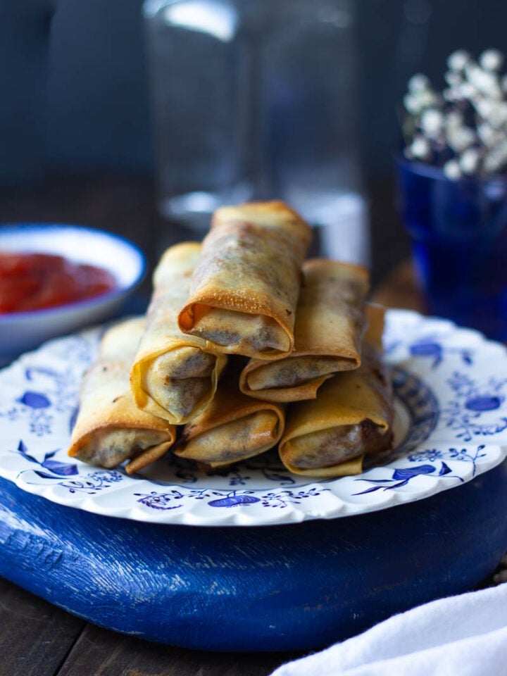 A stack of golden-brown Southwest Rolls is arranged on a decorative blue and white plate. In the background, a small bowl of red dipping sauce and blurred jars sit under soft lighting that highlights the food.