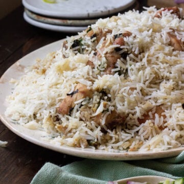A plate of White Biryani with visible pieces of chicken, garnished with herbs and served on a wooden table beside a cucumber and onion salad in a small bowl. Plates and lime slices are in the background.