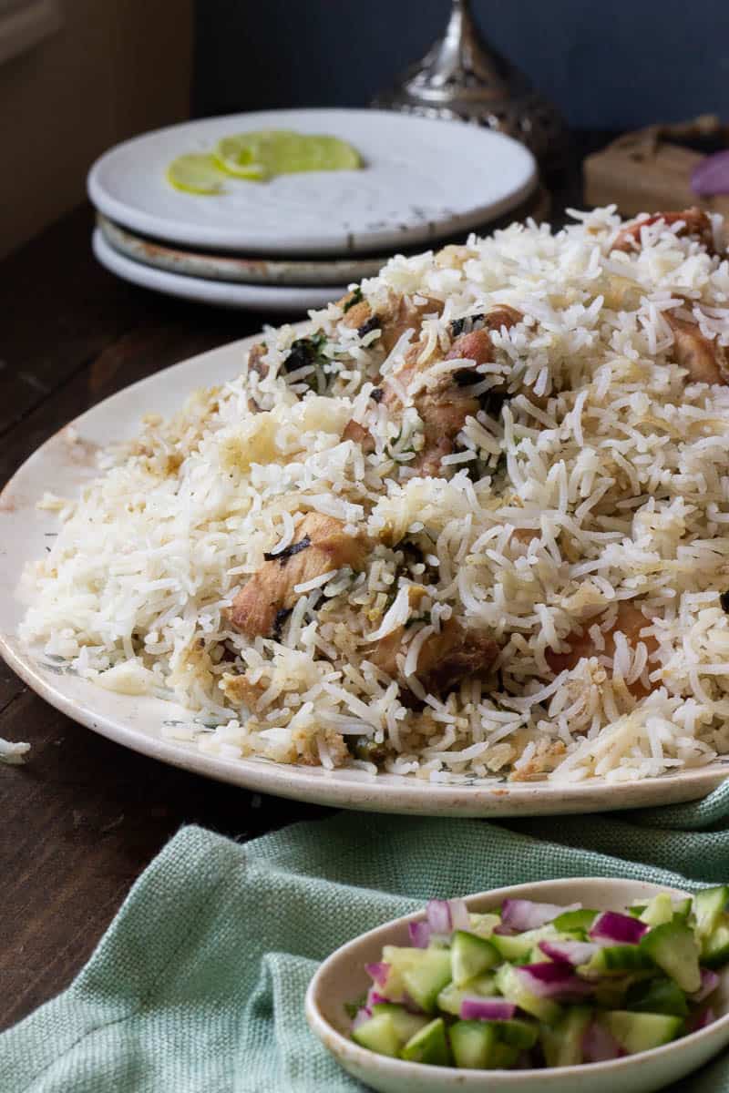 A dish of white biryani in ¾ view. A small dish of cucumbers and onions in the foreground