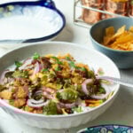 A bowl of Mixed Plate Chaat topped with sliced red onions, cilantro, green and brown chutneys, beside bowls of chickpeas, chips, and garnishes on a white table with decorative dishes in the background.