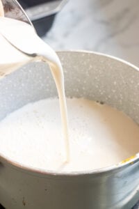 A close-up of milk being poured from a glass measuring cup into a speckled gray saucepan on a kitchen counter, the first step in preparing creamy Caramel Custard.