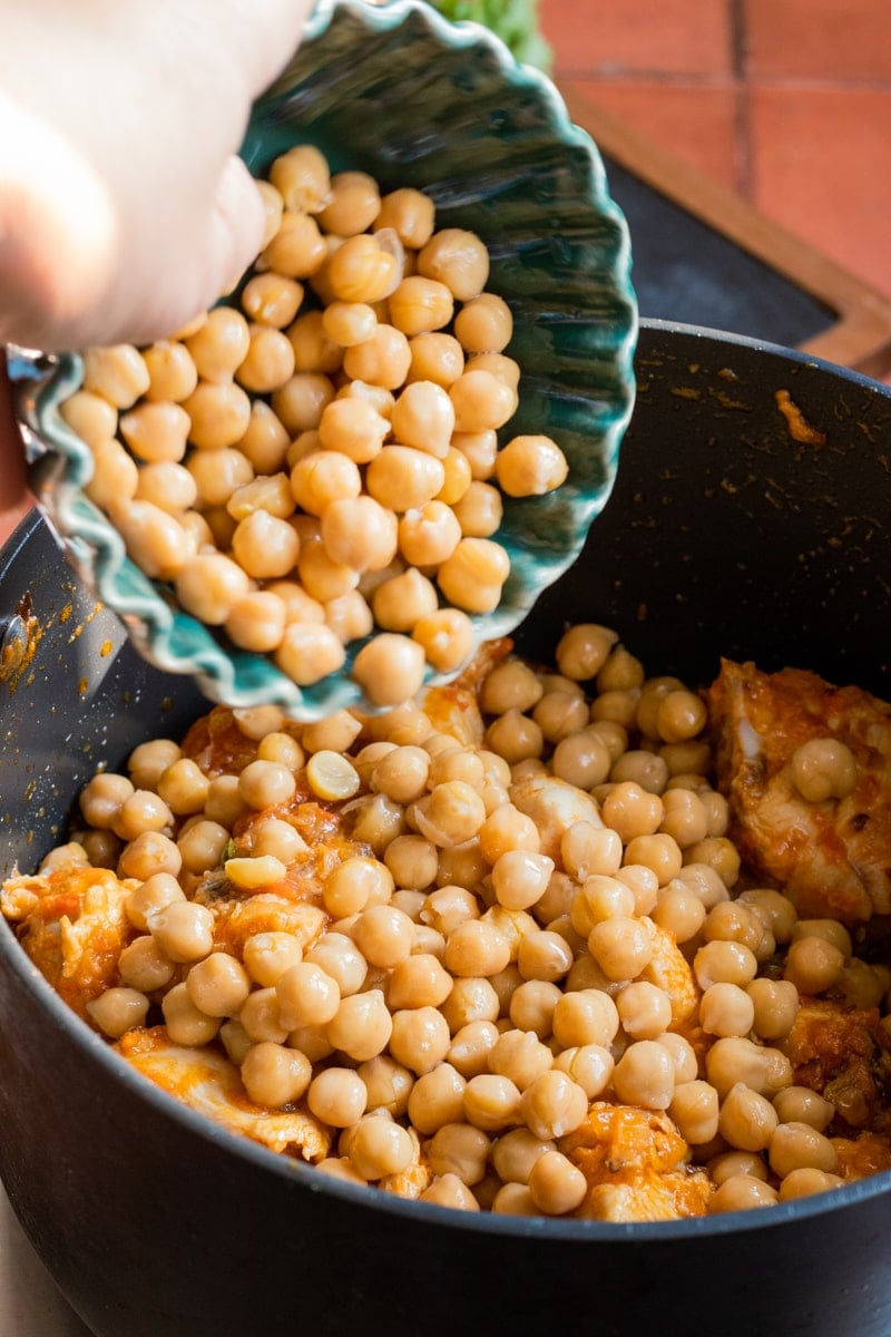 A hand pours chickpeas from a bowl into a pot containing a chunky, orange-colored Chana chicken stew, also known as Murgh Cholay.