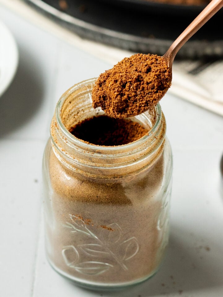 A close-up of a glass jar filled with ground coffee. A spoon is held above the jar, scooping out the coffee powder-reminiscent of how Pakistani Garam Masala is carefully measured. The jar sits on a white surface.