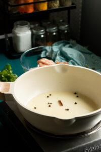A white pot on a stovetop contains pale liquid and whole spices, hinting at white chicken curry in the making. In the background are glass jars, raw chicken, herbs, and a blue cloth.