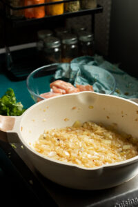Chopped onions caramelizing in a white pan on a stovetop, with raw chicken pieces ready for white chicken curry, spices, and a blue towel visible in the background.