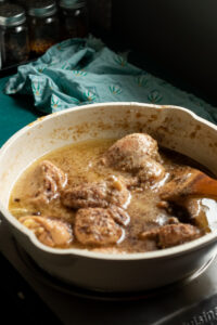 Chicken pieces simmering in a creamy, brownish white chicken curry sauce in a white pot on a stovetop, with a wooden spoon inside, and spice jars and a green cloth in the background.