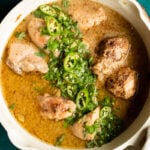 A creamy white chicken curry in a white pan, garnished with chopped cilantro and sliced green chilies, set against a teal background.