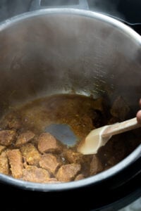 Chunks of beef being cooked for Tala hua gosht in a sauce inside a metal Instant Pot, with a white spatula stirring the meat. Steam rises from the pot, and measurement markings are visible on the pot's inner wall.