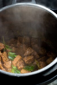 Chunks of cooked beef, known as Tala hua gosht, with spices and fresh green curry leaves inside a steaming metal pressure cooker.