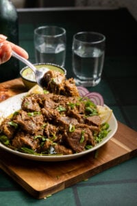 A plate of spicy Tala hua gosht curry garnished with chopped herbs, beside lemon slices and red onion rings, on a wooden board. A fork lifts a piece of meat, with two glasses of water in the background.