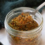 A close-up of a glass jar filled with colorful Achari Masala spice blend. A metal spoon scoops out the mixture, revealing dried herbs and crushed red pepper flakes. A dark cloth is in the background.