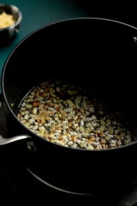 Diced onions being sautéed and caramelized in oil inside a black pot on a stovetop, as the first step in preparing authentic Pakistani Aloo Gobi, with a bowl of minced garlic blurred in the background.