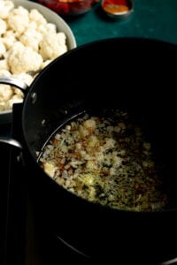 Chopped onions and spices sizzling in oil in a black pot, with a plate of raw cauliflower florets and some spices nearby-a classic start to Pakistani Aloo Gobi.