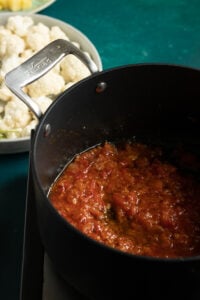 A black pot with tomato sauce simmering inside, placed on a dark surface; in the background, a plate of Pakistani Aloo Gobi cauliflower florets is slightly out of focus.