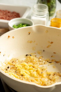 Chopped onions and minced garlic are being sautéed in a light-colored pan for Chicken Keema. Small bowls of chopped green herbs, ground meat, and seasonings are visible in the background.