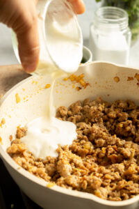 A hand pours a creamy white liquid from a small glass container into a skillet with cooked Chicken Keema and sautéed onions. A jar of salt and green herbs are blurred in the background.