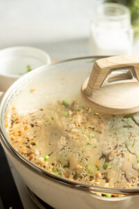 A close-up of a large pot with a glass lid, showing condensation inside and food cooking, possibly Chicken Keema or a grain dish, with green herbs visible. The pot is on a stovetop with a blurry background.