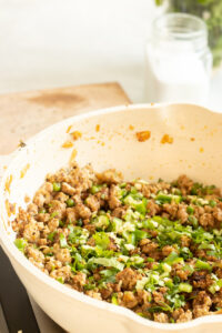A white skillet filled with flavorful Chicken Keema, topped with chopped green onions and fresh cilantro, sits on a stovetop. A glass jar and a cutting board are blurred in the background.