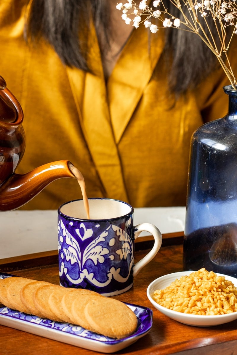 tea time set up: a wooden tray with a mug, some snacks. tea being poured from a kettle into a mug. person wearing yellow in the background 