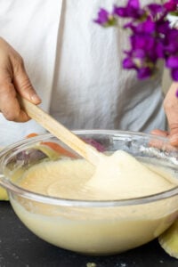 A person in a white shirt stirs thick Marble Cake with Oil batter in a clear glass bowl with a wooden spoon; purple flowers are blurred in the background.