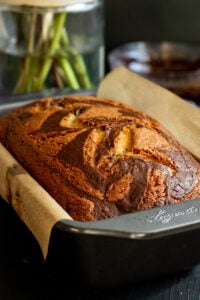 A Marble Cake with Oil, featuring swirls of chocolate and vanilla, rests in a loaf pan lined with parchment paper on a dark surface, with a blurred vase and bowl in the background.