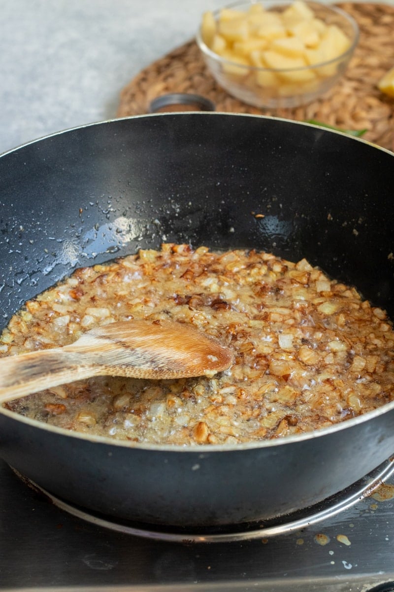 chopped onions and ginger and garlic being sauted in a black wok with a wooden spoon