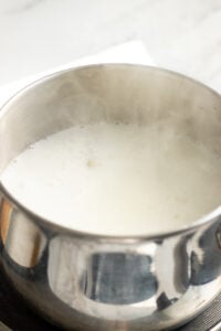 A stainless steel pot filled with boiling milk, frothy and bubbling at the top, is perfect for making Doodh Patti. Steam rises from the surface as it heats on the stove, capturing the essence of this classic tea preparation.