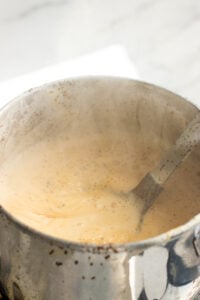 A close-up of a metal pot filled with frothy, bubbling Doodh Patti chai, with steam rising and a spoon stirring the creamy mixture. The background is bright and slightly out of focus.