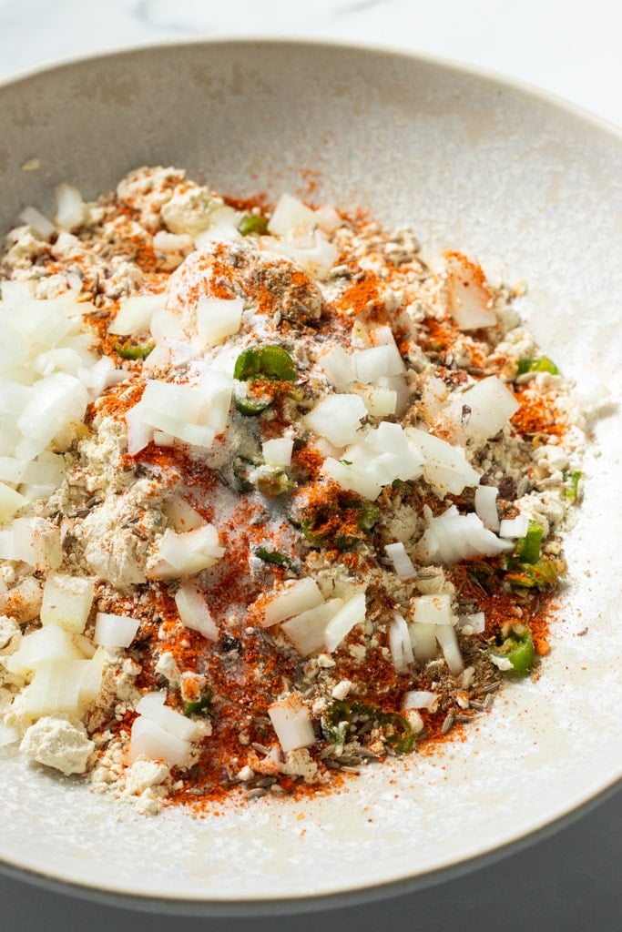 A close-up of a bowl containing chopped onions, green chilies, assorted spices, and flour mixture, ready to be mixed for a flavorful Kadhi Pakora recipe.