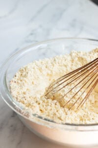 A glass bowl filled with pakora mix sits on a marble surface, with a metal whisk resting inside the bowl-an essential first step in learning how to make pakoras.