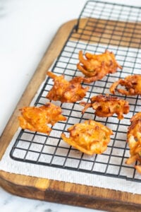 Golden-brown vegetable fritters made with pakora mix rest on a wire cooling rack over a paper towel on a wooden tray.