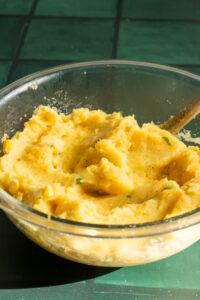 A glass bowl filled with creamy mashed potatoes, perfect for making Aloo Cutlets, is garnished with herbs and sits on a green tiled surface. A wooden spoon is partially submerged in the mashed potatoes.
