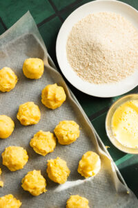 Spoonfuls of yellow dough for this Potato Cutlets Recipe are arranged on a parchment-lined tray beside a plate of breadcrumbs and a bowl of beaten egg, ready for breading on a green tiled surface.