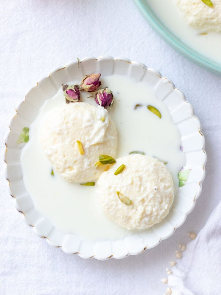 two pieces of soft milk powder rasmalai on a plate with some dried roses