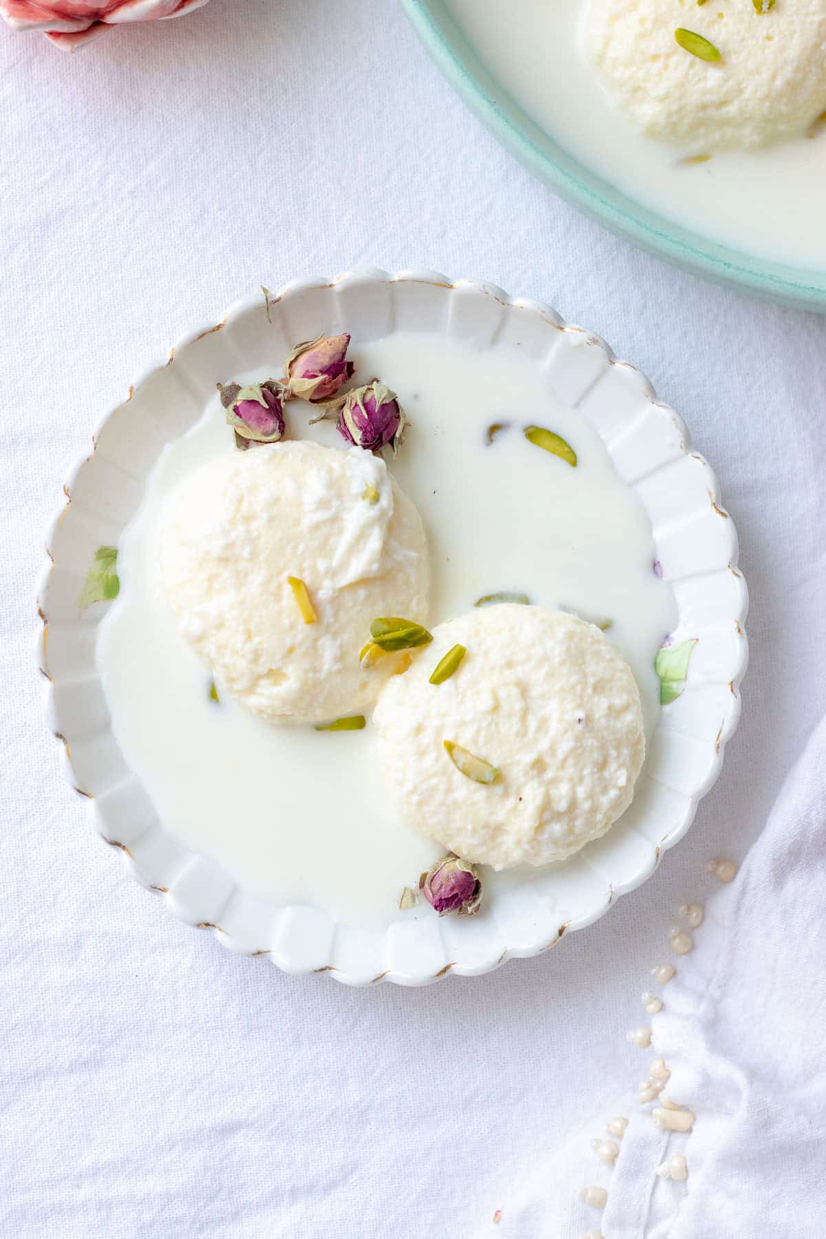 2 ras malai pieces in a plate with milk, a dried rose and pistachios for garnish