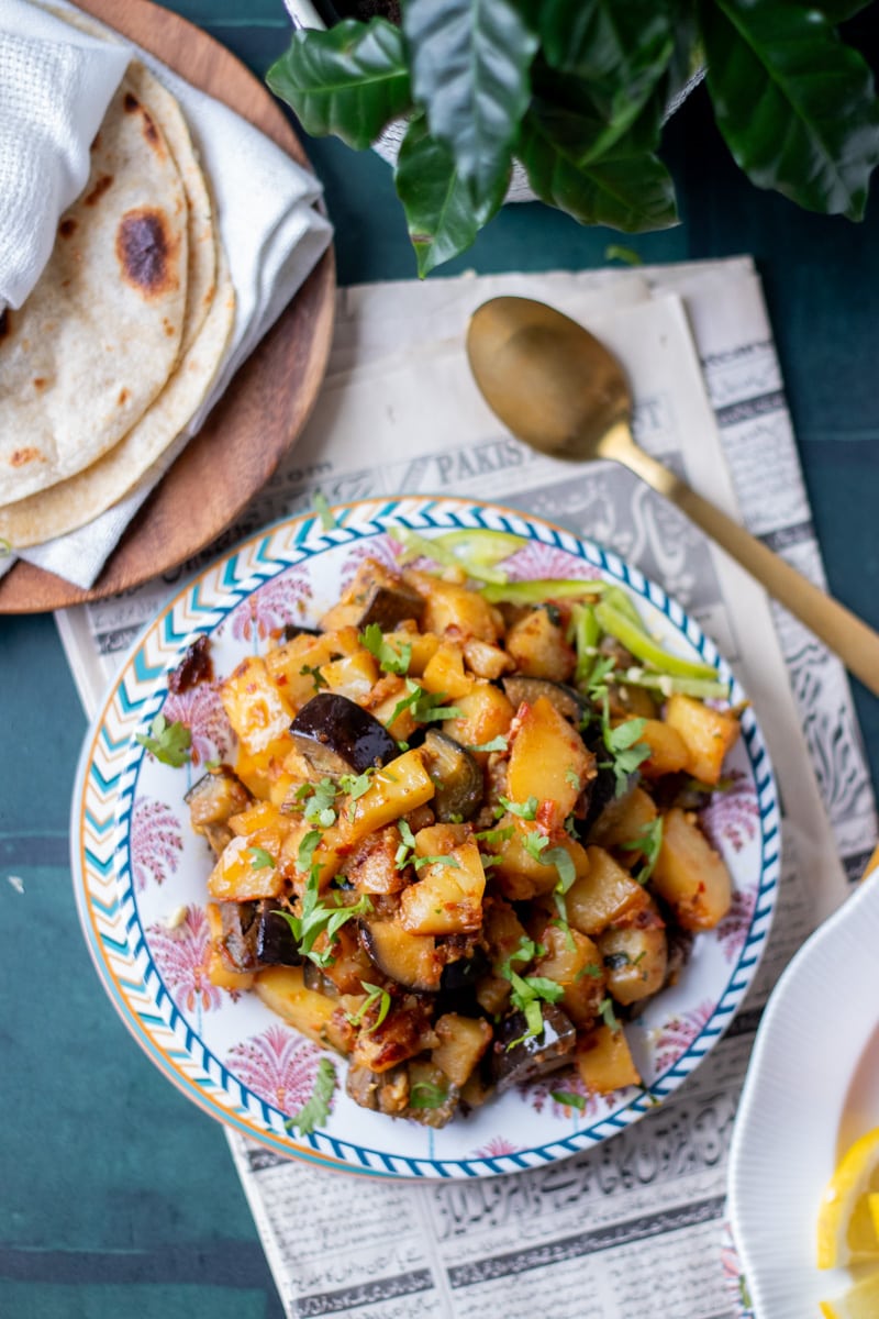 Aloo Baingan in a dish, chapati in a plate in the corner, a gold spoon on the side and a plant in the top