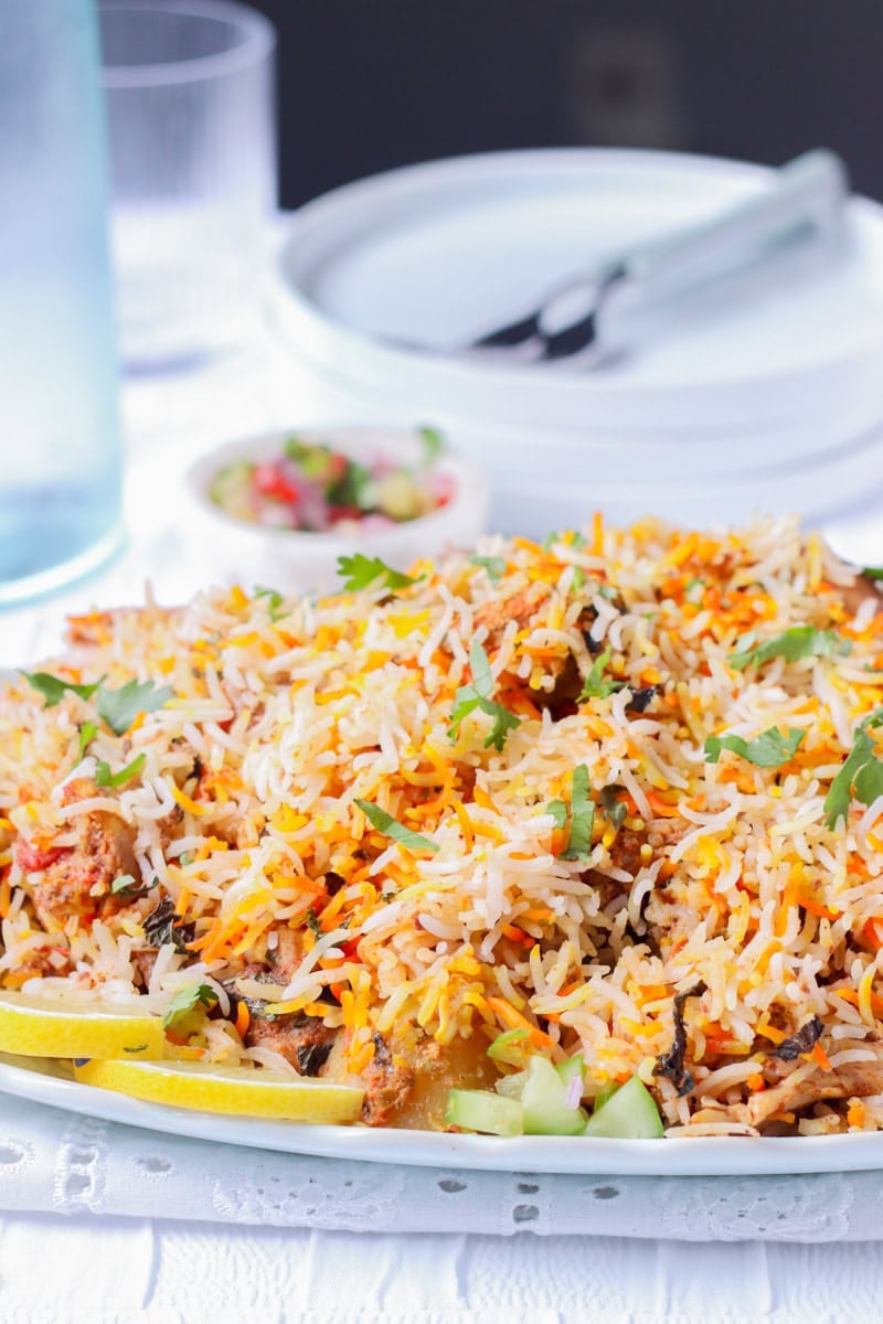 Chicken Biryani on a platter, plates, a water bottle, a glass and a small bowl of kachumbar in the background