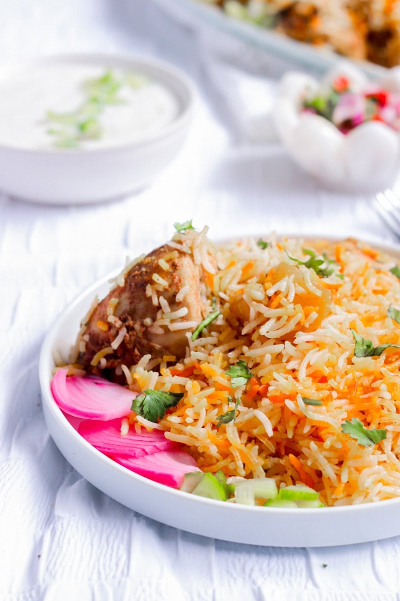 chicken biryani in a plate with pickled onions and cucumbers. a bowl of raita and kachumbar in the back